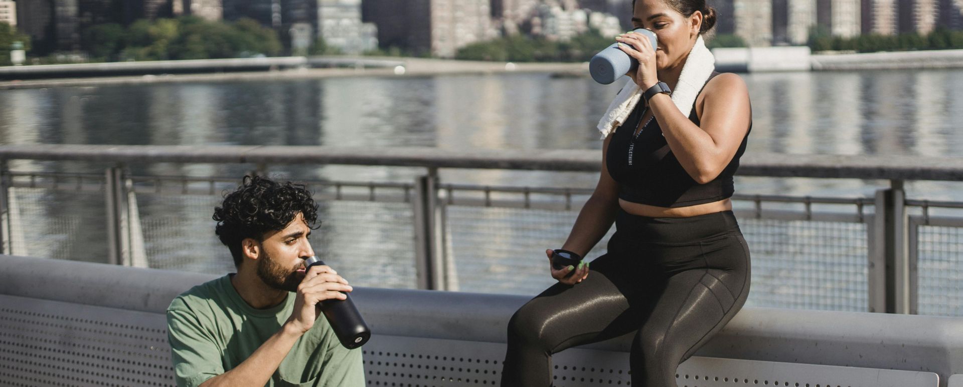 A young couple takes a break by the waterfront to hydrate after exercising outdoors in a sunny city setting.