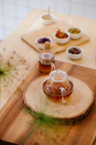 A glass teapot on a round wooden board surrounded by various tea ingredients in small bowls.