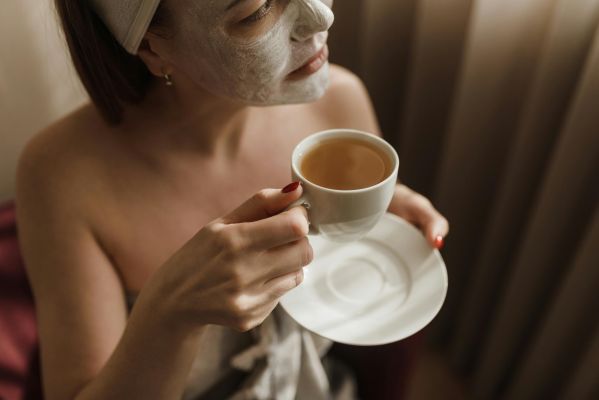 A woman with a facial mask enjoys a warm beverage during a skincare routine.