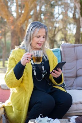 Elderly woman sitting outdoors, drinking tea and using a smartphone in a sunny setting.